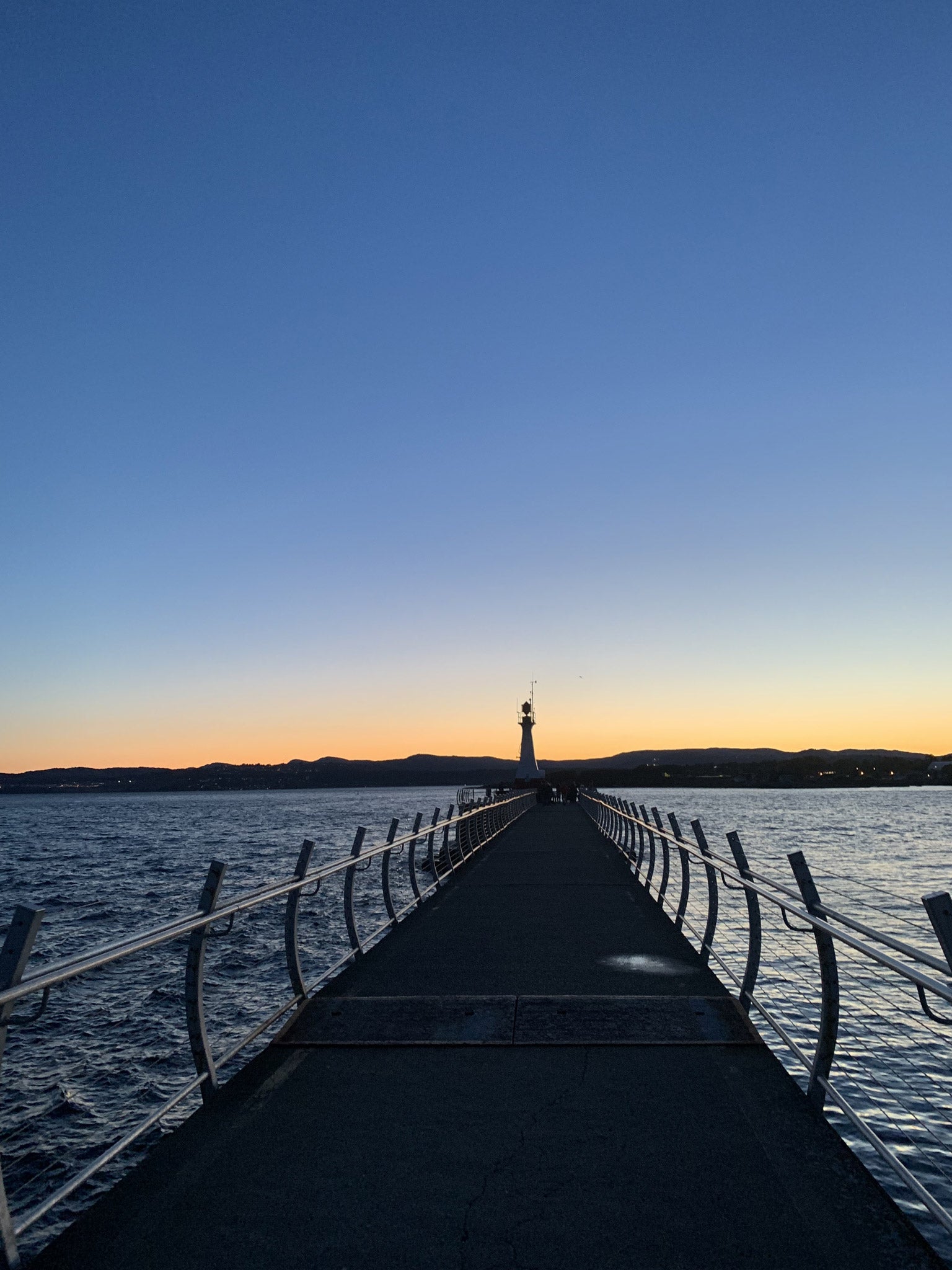 Pier extending into a body of water with a lighthouse in the distance at sunset at Odgen Point in Victoria, BC